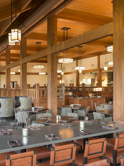 The image shows an empty, modern dining area with wooden beams, tables set with cutlery, and upholstered chairs in a bright, spacious setting.