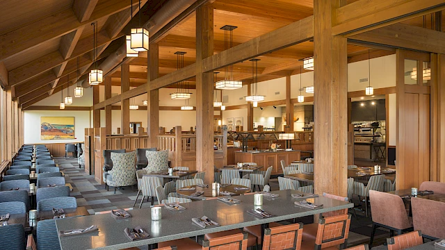 The image shows an empty, modern dining area with wooden beams, tables set with cutlery, and upholstered chairs in a bright, spacious setting.