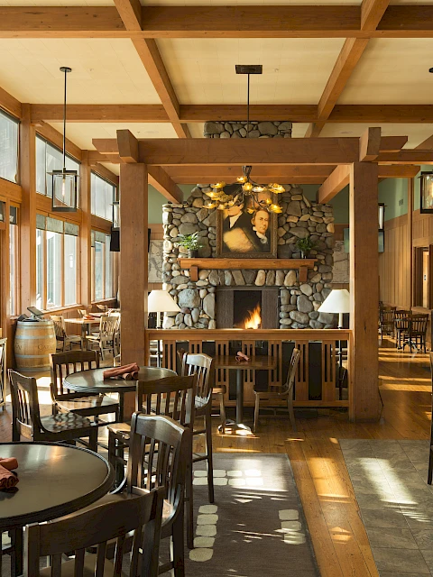 A cozy restaurant with wooden furniture, a stone fireplace, and large windows letting in natural light.