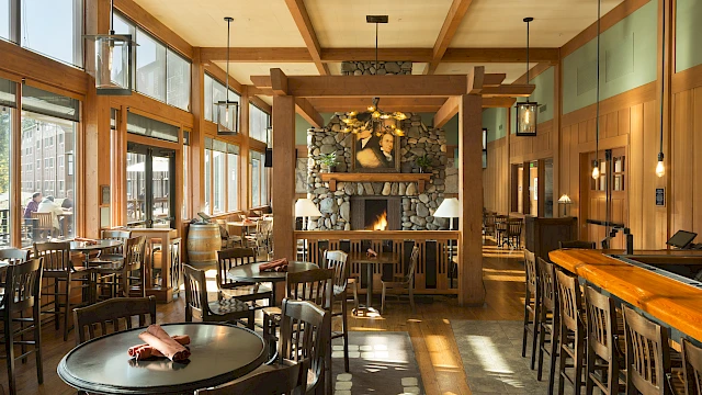 A cozy restaurant with wooden furniture, a stone fireplace, and large windows letting in natural light.