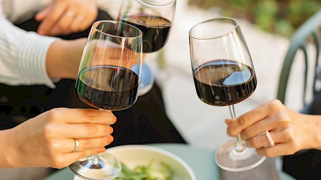 Three people are clinking glasses of red wine over a table with a salad.