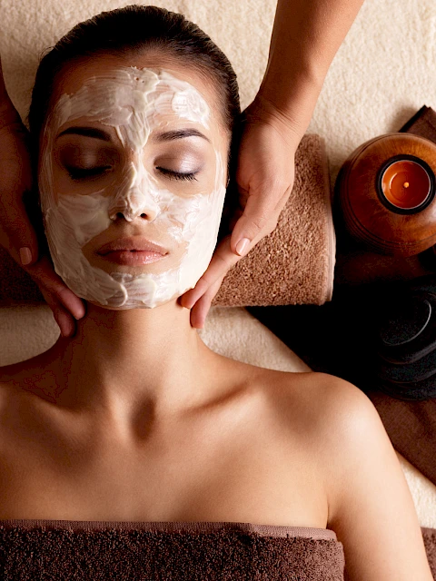 A person receiving a facial treatment with a mask applied, lying on a towel, with candles and massage stones nearby.