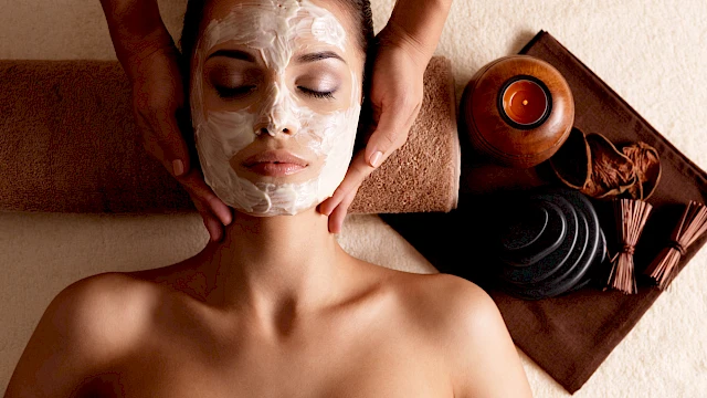A person receiving a facial treatment with a mask applied, lying on a towel, with candles and massage stones nearby.
