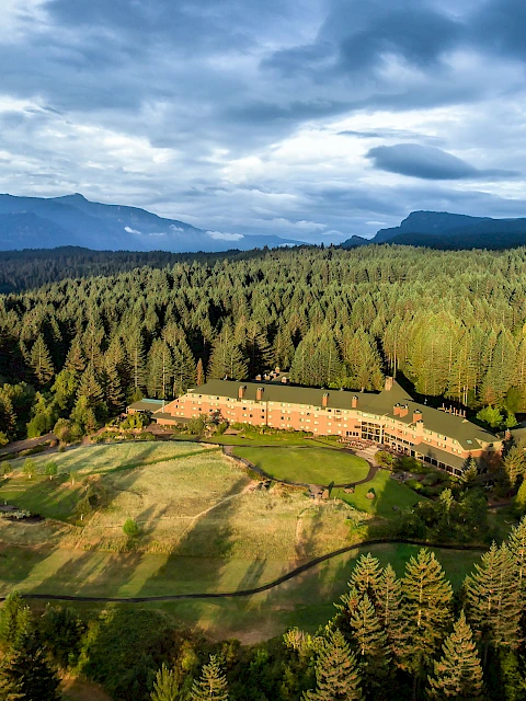 A large building surrounded by dense forest, mountains, and a body of water under a cloudy sky.