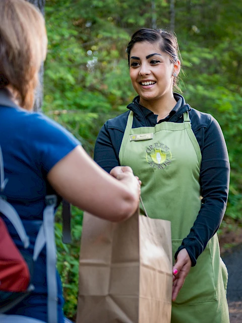 A woman in a green apron is handing a paper bag to another person in an outdoor setting.
