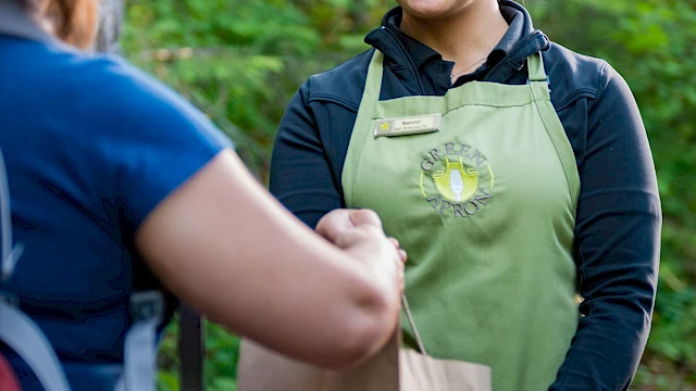 A woman in a green apron is handing a paper bag to another person in an outdoor setting.