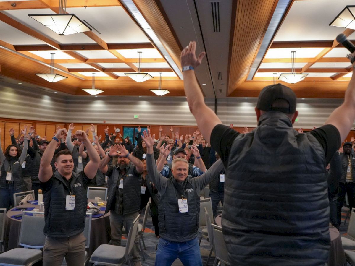 A person on stage leads an enthusiastic crowd, raising arms, in a conference room setting with ceiling lights and tables around.