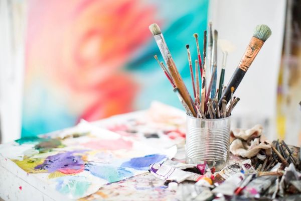 Paintbrushes in a tin can and tubes of paint are on a table with a colorful abstract painting blurred in the background.