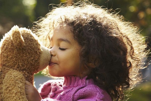 A child with curly hair kisses a teddy bear while wearing a purple sweater in a sunny outdoor setting.