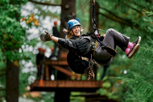 A person wearing a helmet and safety gear is zip-lining through a forest, smiling and giving a thumbs-up, with trees in the background.