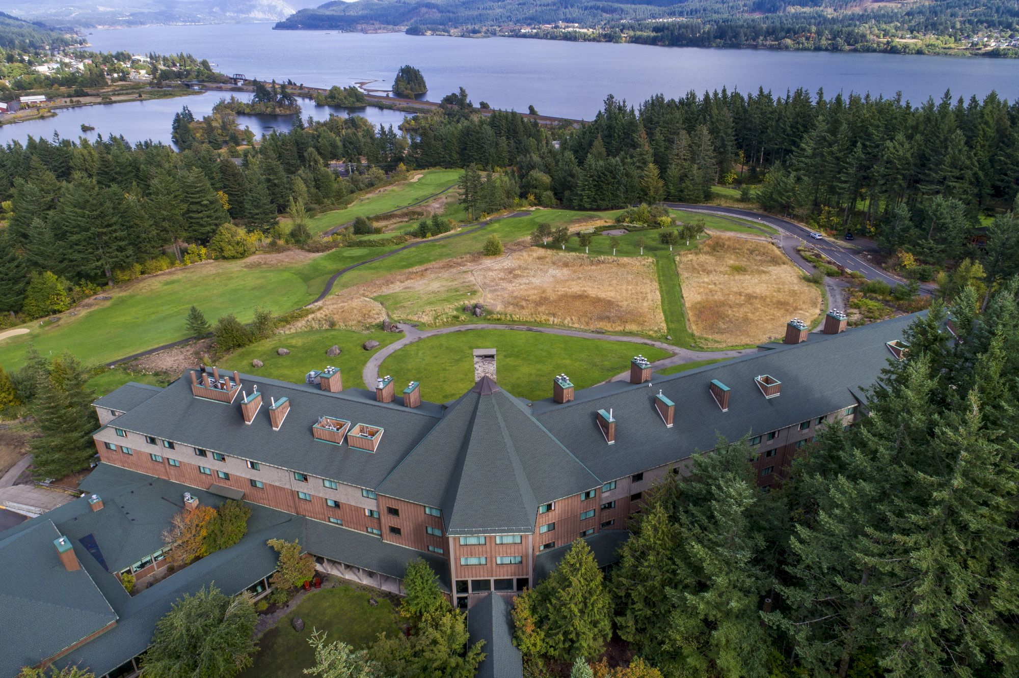 An aerial view of a large building surrounded by lush greenery, forest, and a river or lake in the background.
