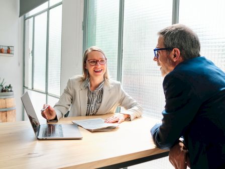 Two people are sitting at a table in an office, discussing something with a laptop and documents in front of them.