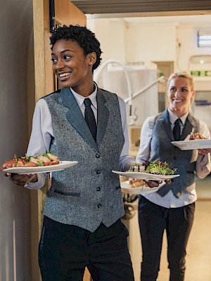 Two smiling servers in vests and ties carry plates of food through a doorway, likely in a restaurant or catering setting.