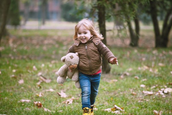 A smiling child in a brown jacket walks through a grassy area while holding a teddy bear. Leaves are scattered on the ground.