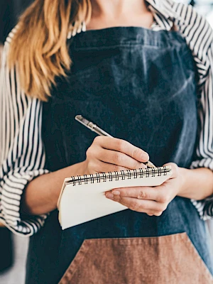 A person wearing an apron is holding a notepad and pen, possibly taking orders in a restaurant.