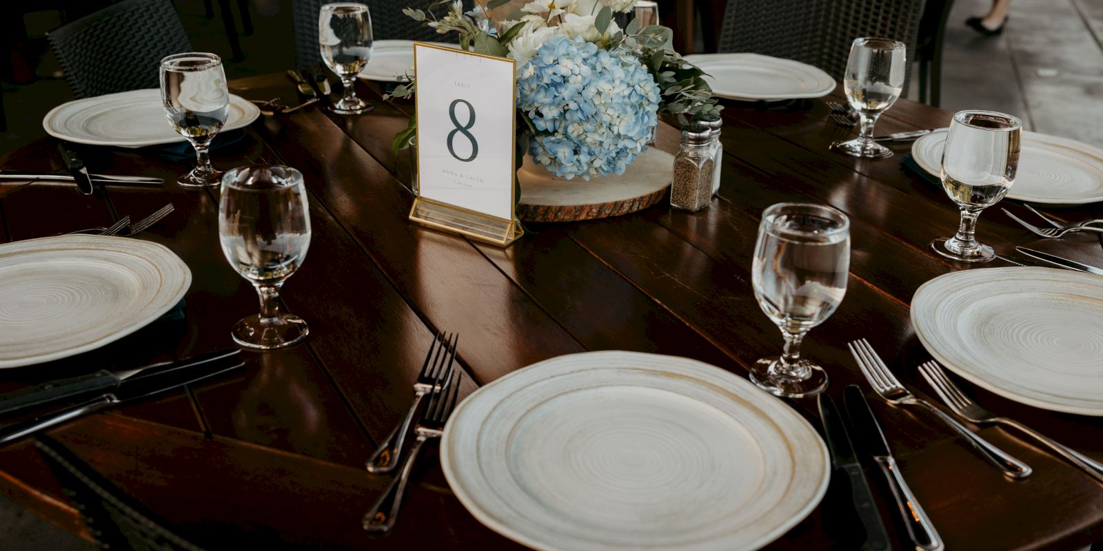 A table is set with six place settings, glasses, cutlery, and a centerpiece with blue flowers, marked with the number 8.
