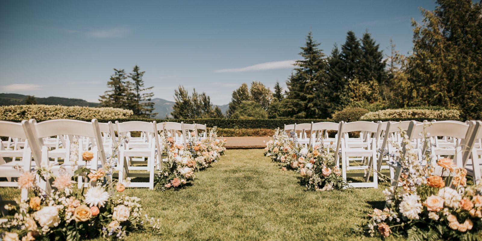 An outdoor wedding setup features rows of white chairs and floral arrangements on grass, surrounded by trees and a clear blue sky.