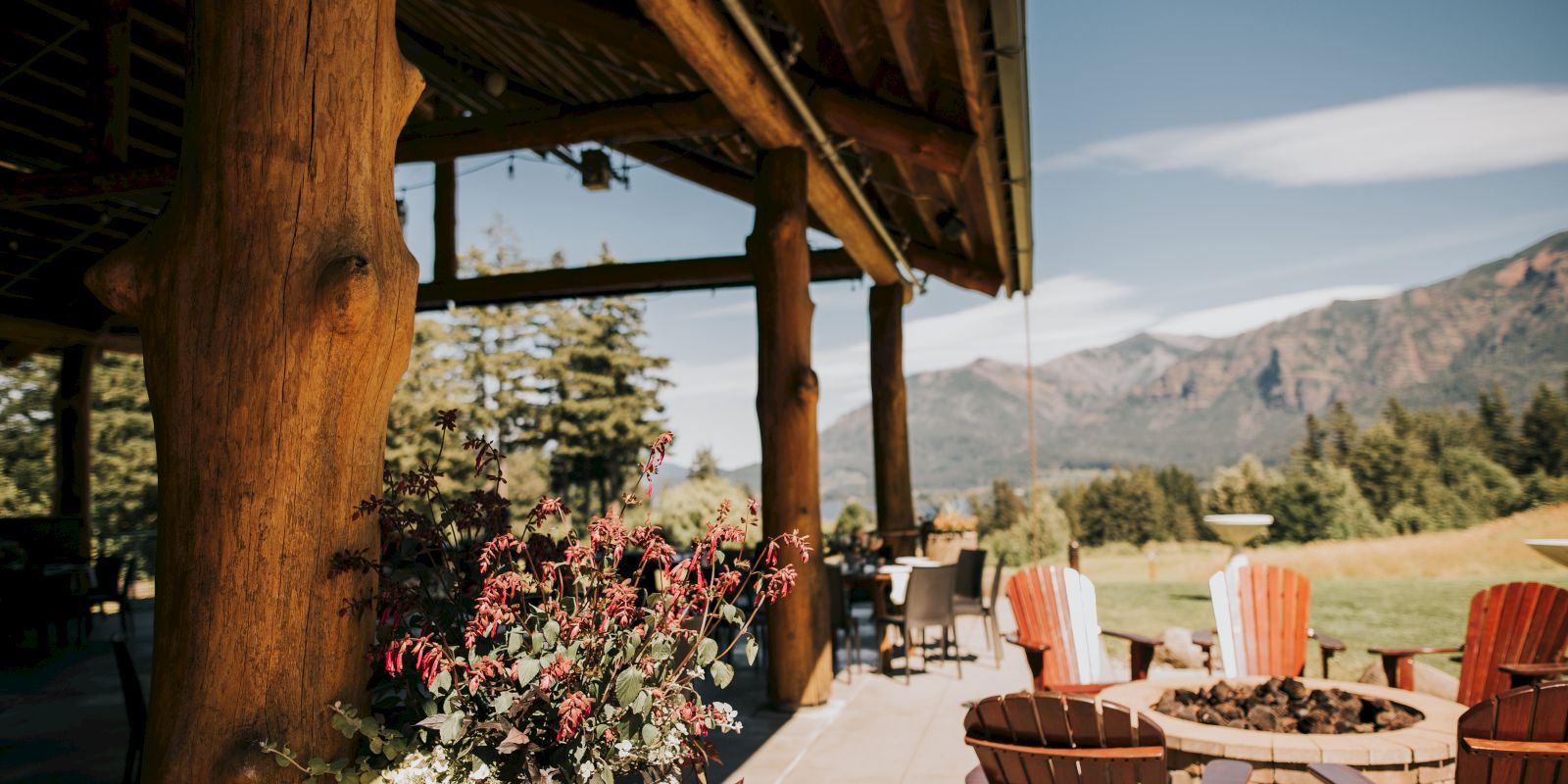 A rustic patio features wooden beams, Adirondack chairs around a fire pit, potted flowers, and a scenic mountain view in the background.