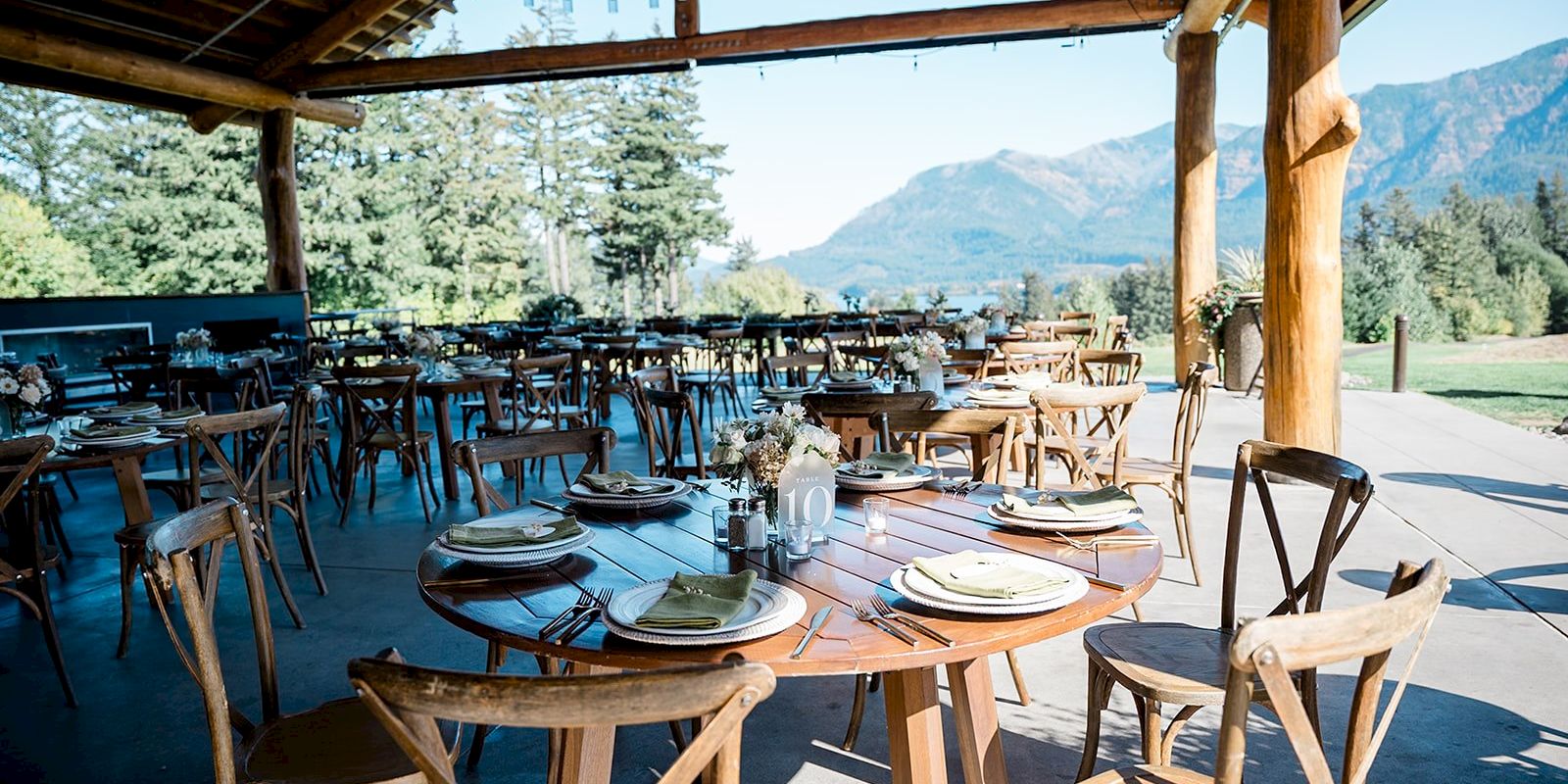 An outdoor wedding reception setup with wooden tables and chairs, set against a backdrop of trees and mountains under a wooden pavilion.