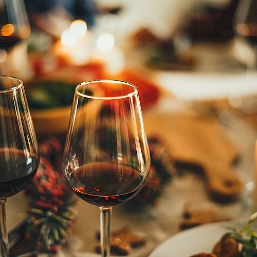Two glasses of red wine are on a table with food, candles, and festive decorations, creating a warm, cozy atmosphere.