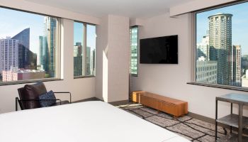 A modern hotel room with large windows showing tall city buildings, a wall-mounted TV, armchair, and bed are seen.