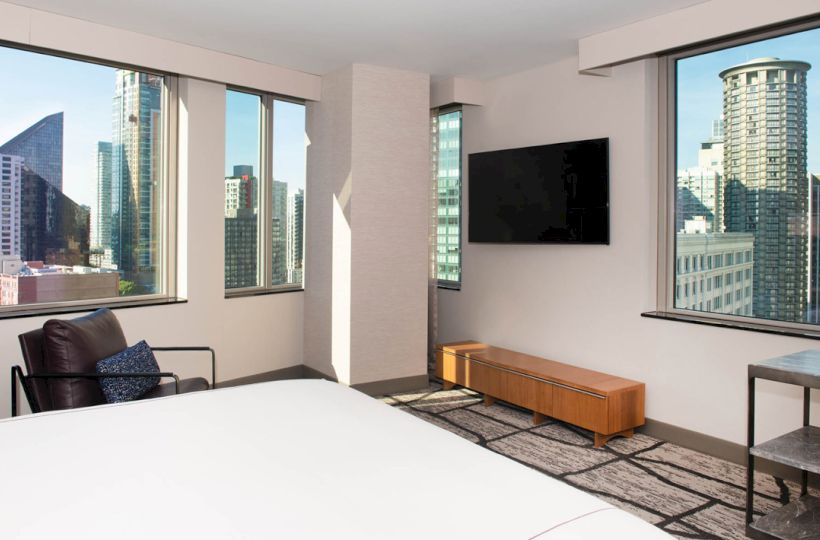 A modern hotel room with large windows showing tall city buildings, a wall-mounted TV, armchair, and bed are seen.