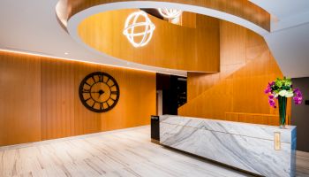 A modern lobby with a wooden spiral design, wall clock, marble reception desk, and flower vase.