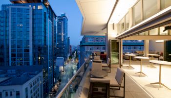 A modern cityscape at dusk with tall buildings, viewed from a balcony that has tables and chairs under bright lighting.