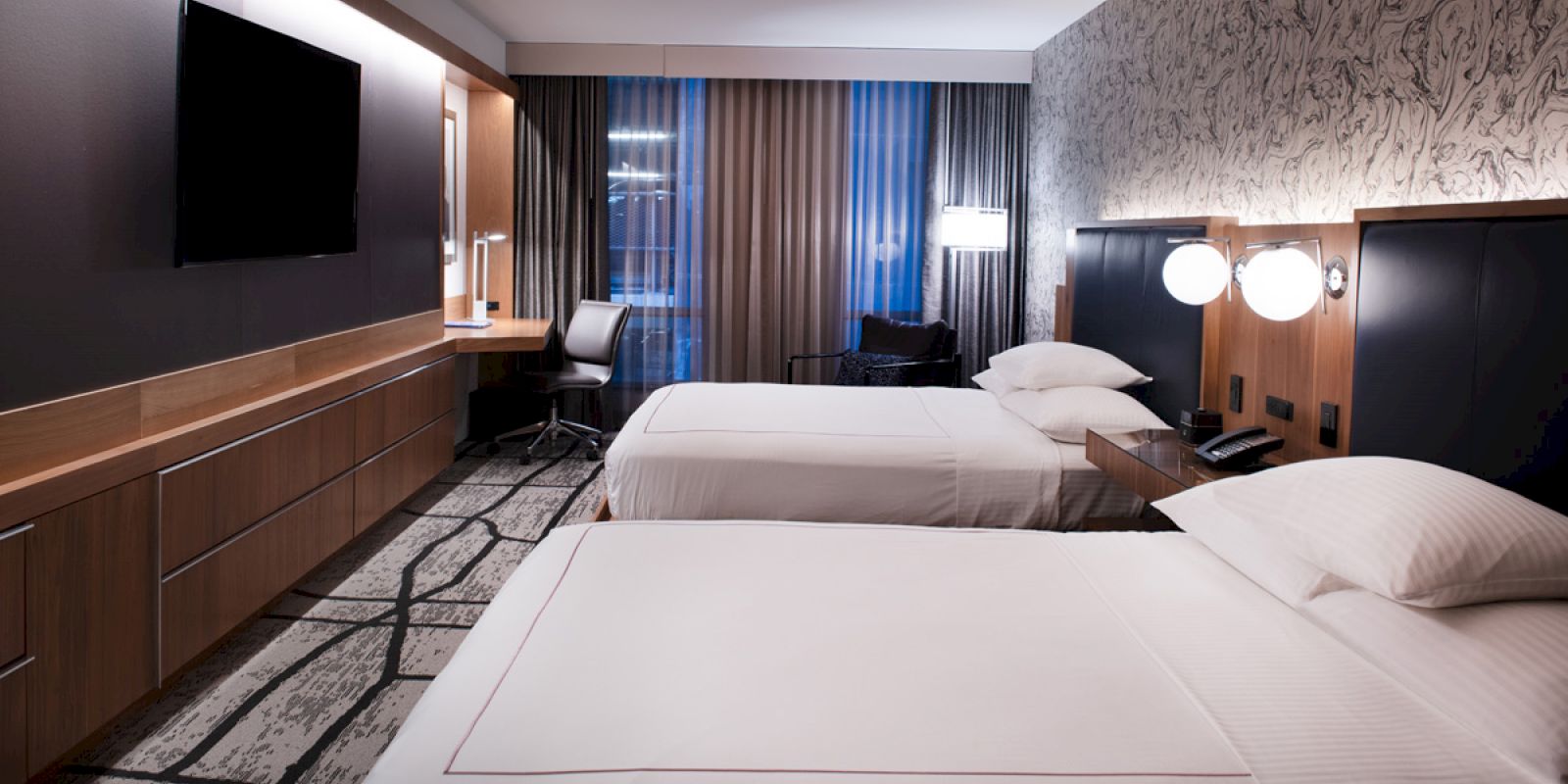 The image shows a modern hotel room with two beds, a large TV, a desk, and a window with closed curtains.