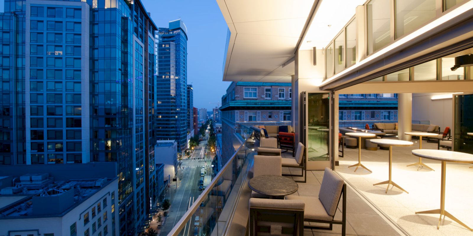 A modern balcony with outdoor seating overlooks a city street, surrounded by tall buildings during evening.