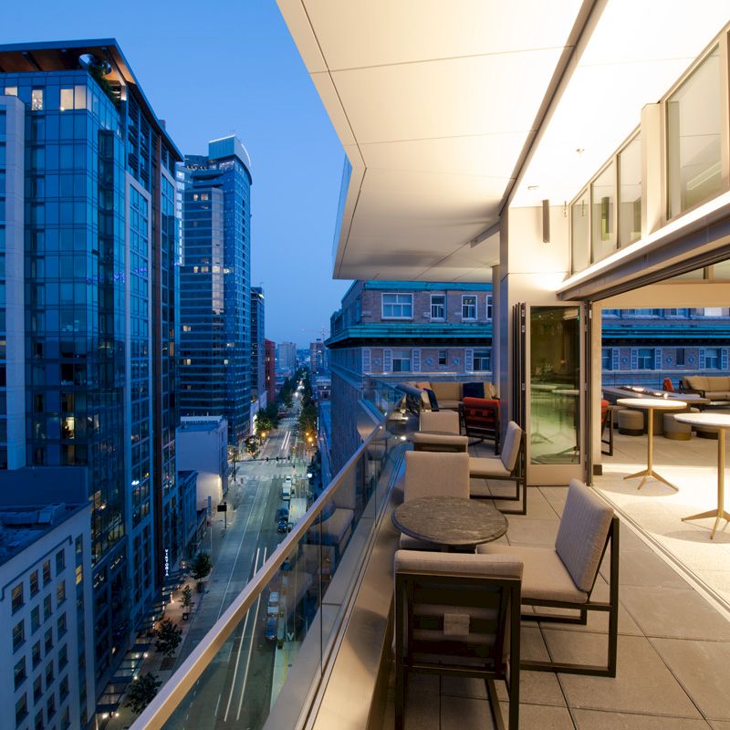 A modern high-rise balcony overlooking a city skyline at dusk, featuring outdoor seating and tables, with buildings lit up.