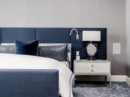 Modern bedroom with a blue upholstered headboard, white bedding, blue accents, nightstand with a lamp, and a decorative piece.
