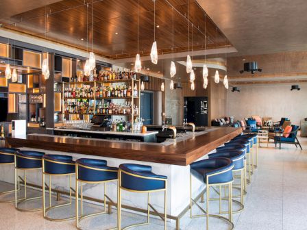 A modern bar with blue stools, a wide selection of bottles, stylish lighting, and a cozy seating area.