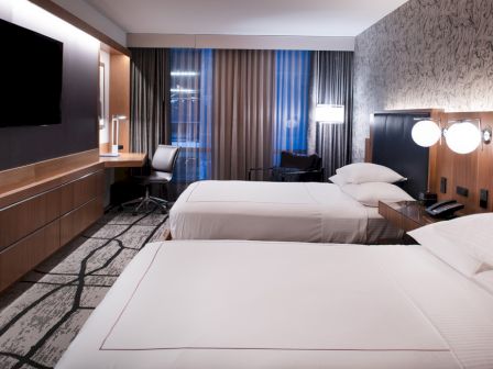 The image shows a modern hotel room with two neatly made beds, a desk with a chair, a mounted TV, and stylish lighting.