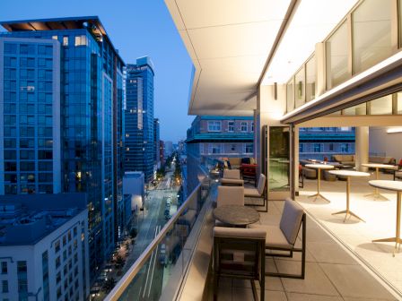 A modern rooftop terrace with seating overlooks a cityscape of tall buildings at dusk, featuring a lit interior space.