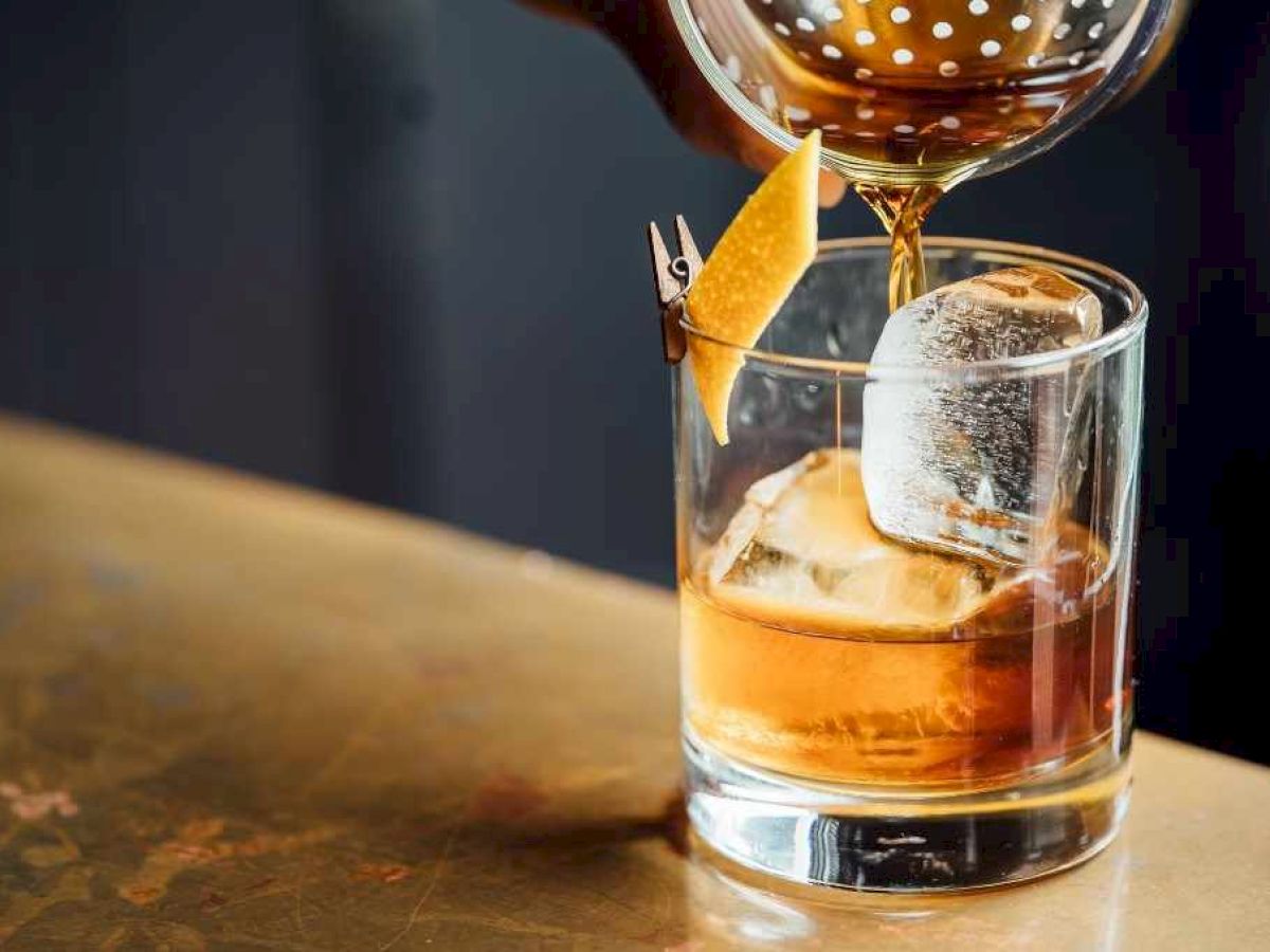 A hand pours a brown liquid from a strainer into a glass with ice and garnished with a twist of lemon on a wooden surface.