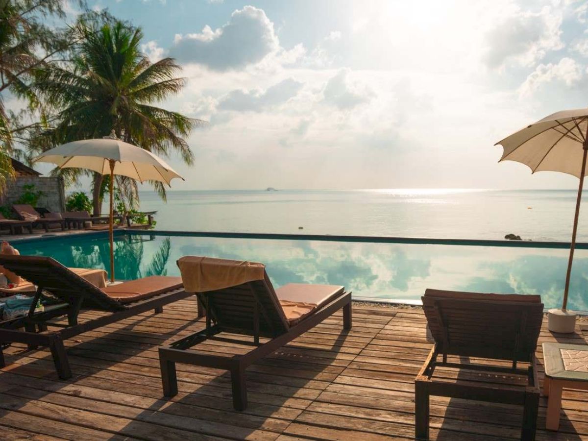 Poolside view with lounge chairs, umbrellas, and palm trees facing a serene ocean under a partly cloudy sky.
