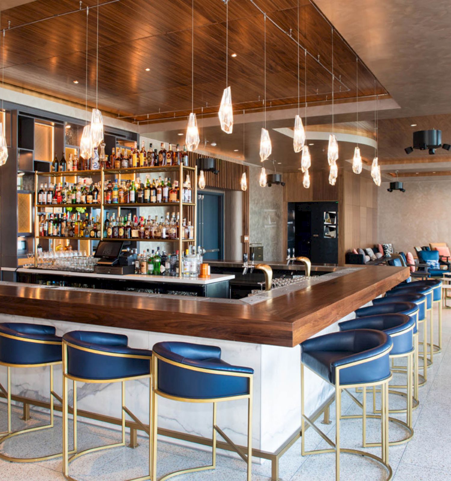 A modern bar with blue stools, hanging lights, and a fully stocked shelf; seating area visible in the back, featuring a stylish design.