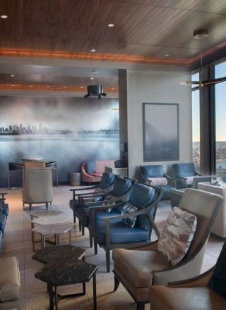A modern lounge with armchairs, small tables, a wall mural, and large windows offering a view of the sea and a distant Ferris wheel.