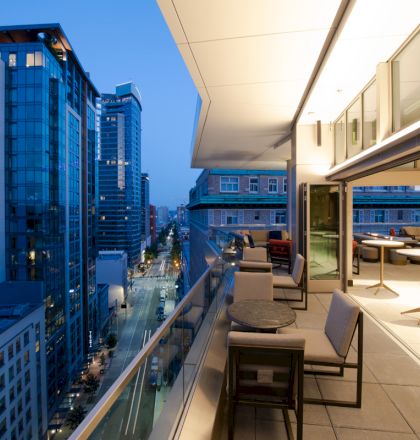 A modern balcony overlooks an urban street with high-rise buildings at dusk; outdoor seating and tables are visible.