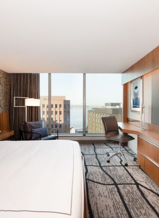 A modern hotel room with a large bed, wall-mounted TV, desk, chair, and a window with a city view.