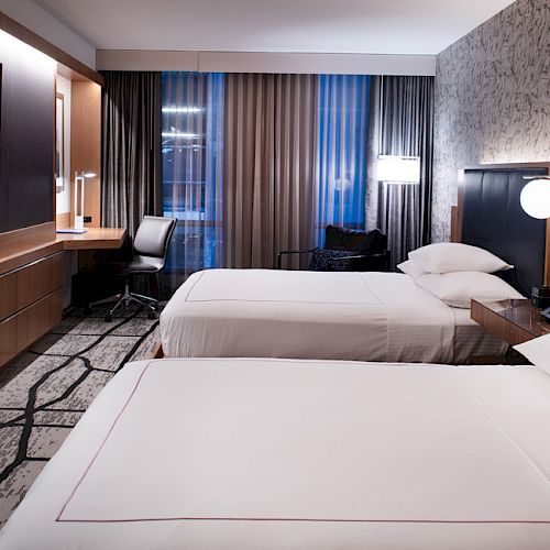 The image shows a modern hotel room with two beds, a TV, a desk, and stylish decor, including patterned wallpaper and carpeting.