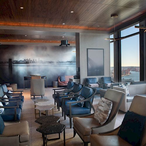 A modern lounge area with chairs and tables, featuring large windows with a scenic water view and a cityscape mural on the wall.