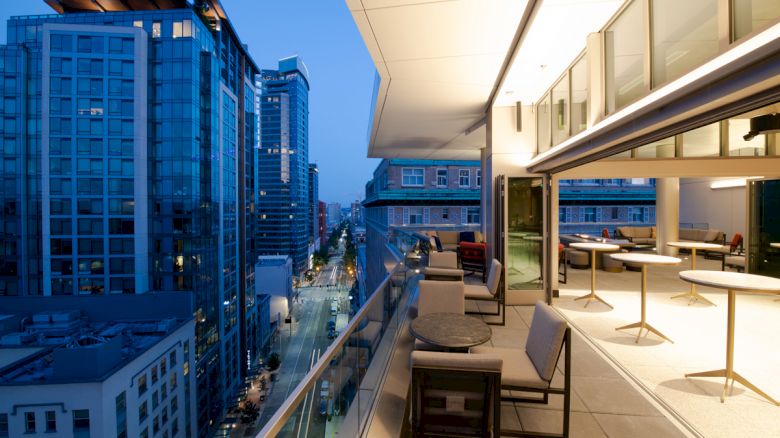 A modern urban balcony with seating overlooks a city street lined with tall buildings, captured at twilight.