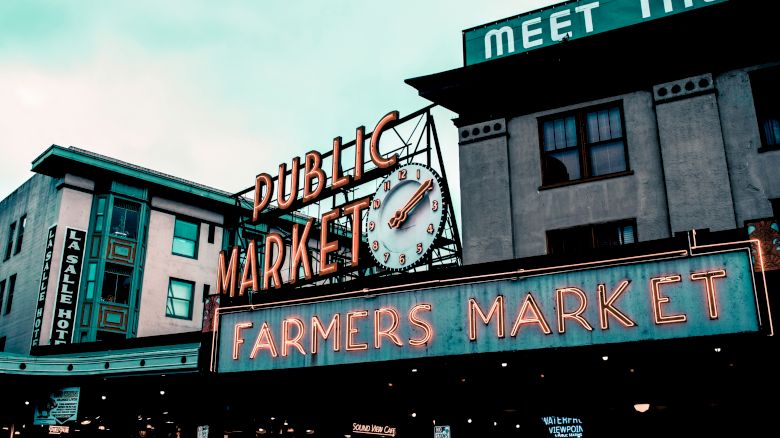 The image shows an iconic farmers market sign with a clock, set against a backdrop of urban buildings under a cloudy sky.