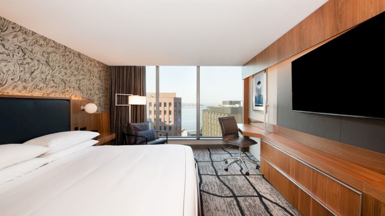The image shows a modern hotel room with a large bed, a desk, a chair, and a view of city buildings from a window.