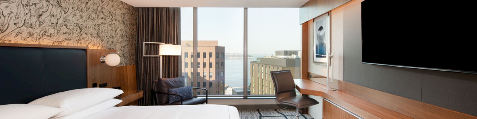 A modern hotel room with a large bed, desk, chair, and a flat-screen TV on the wall, featuring a city view through large windows.
